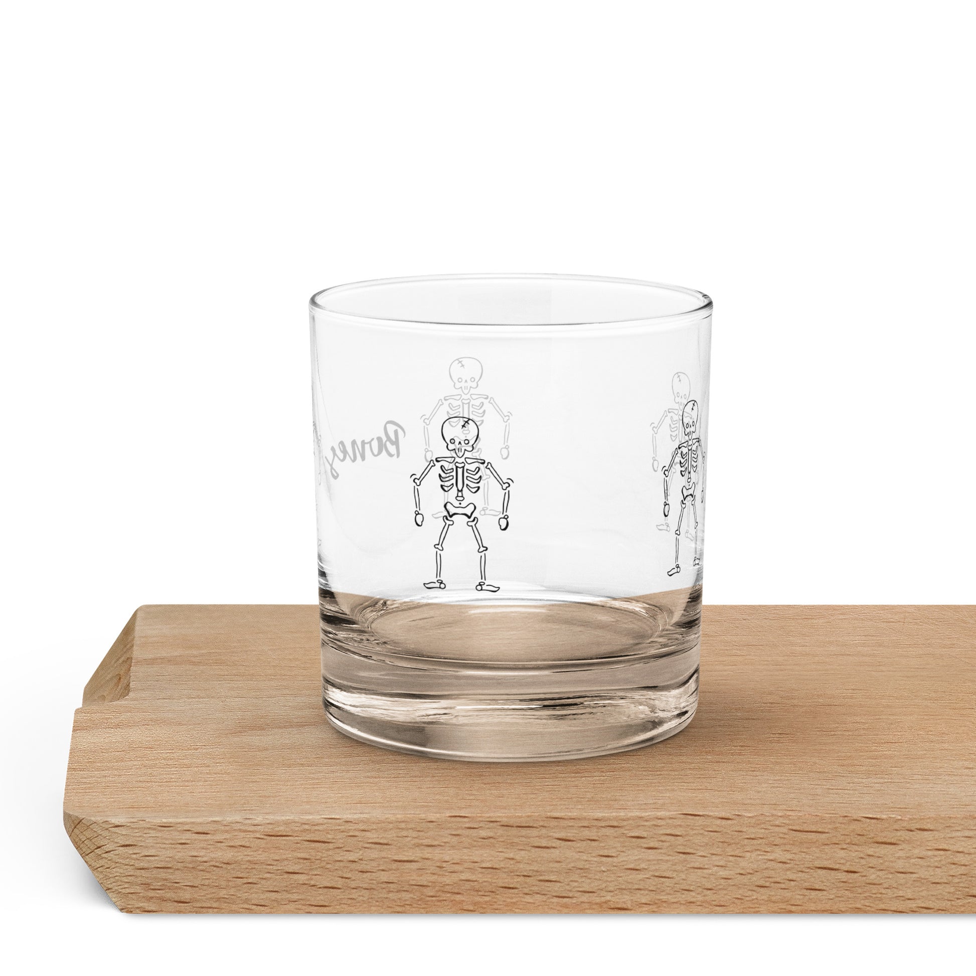 Clear glass rocks glass with black skeleton illustrations and humorous Halloween text on wooden surface.